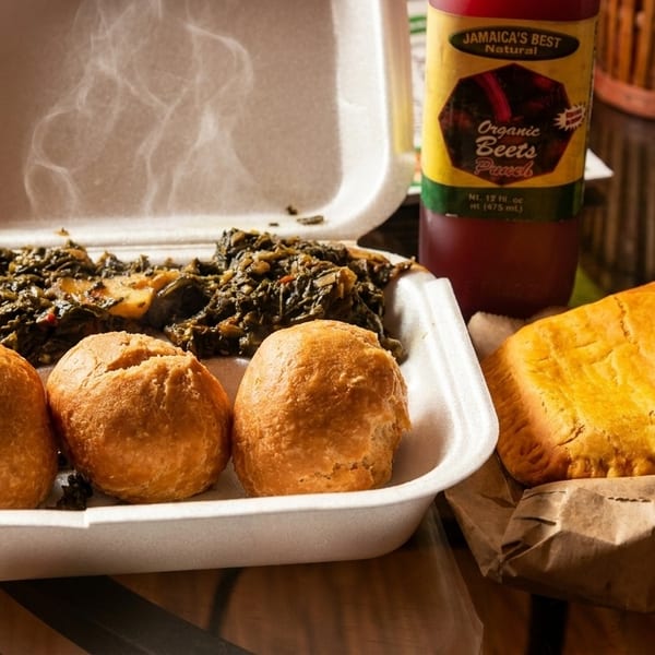 Steaming Jamaican callaloo and dumplings in a takeout container, with a beef patty and beet punch.