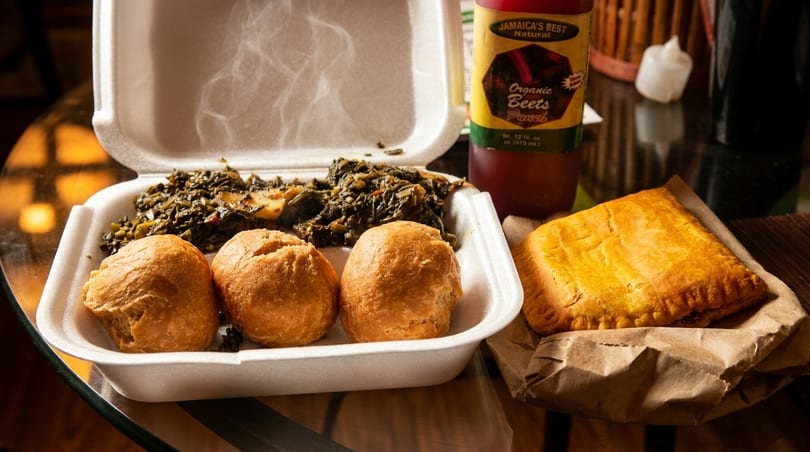 Steaming Jamaican callaloo and dumplings in a takeout container, with a beef patty and beet punch.