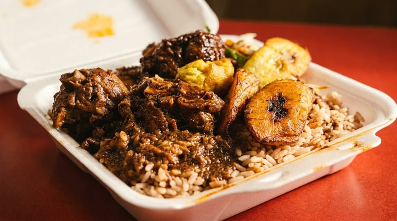 A takeout container filled with brown stew chicken, rice and peas, and fried plantains.