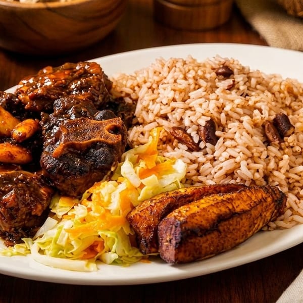 Oxtail stew with rice and peas, fried plantains, and cabbage on a white plate.