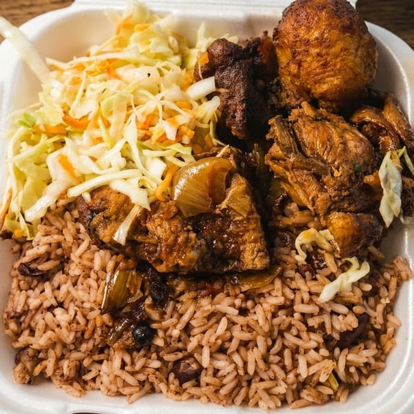 A takeout container filled with rice and peas, brown stew chicken, and coleslaw on a wooden table.